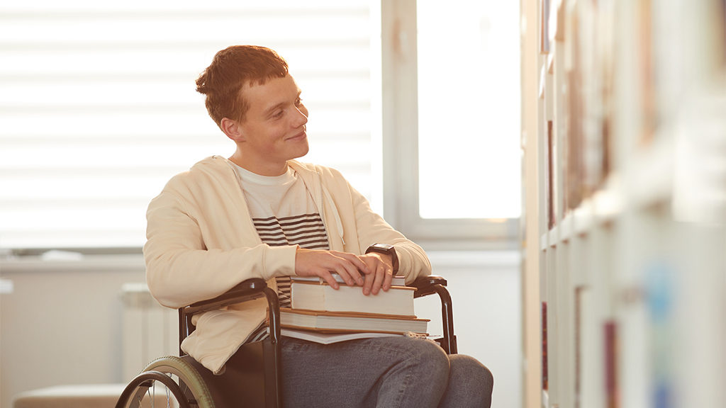 młodzieniec na wózku inwalidzkim patrzący na półki z książkami w bibliotece oświetlonej światłem słonecznym